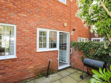 A small patio area with a black barbecue grill next to a brick wall with windows and a glass door surrounded by green plants at Sea View Cottage in Menai Bridge