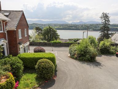 A view of a river with trees and houses along the shore seen from a driveway with bushes and a brick house in Sea View Cottage Menai Bridge