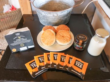 A table with a plate of four bread rolls a jar of jam a bottle of milk packets of Cornish tea and a box of Cornish clotted cream at Honey @ Stars & Embers Glamping in Grampound