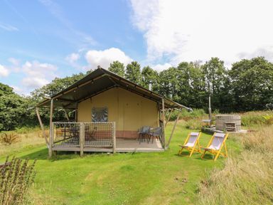 A glamping tent with a wooden deck, outdoor chairs, and a hot tub in a grassy area with trees at Honey @ Stars & Embers Glamping in Grampound