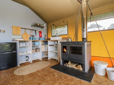 A kitchen area with a black refrigerator a wood-burning stove with a chimney shelves with kitchenware and utensils and a beige rug on the floor at Honey @ Stars & Embers Glamping in Grampound