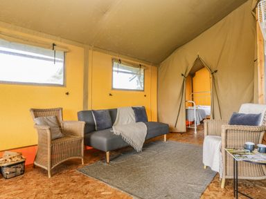 A living area in a tent with wicker chairs, a gray sofa, a rug, and a view into a bedroom with a metal bed frame at Honey @ Stars & Embers Glamping in Grampound
