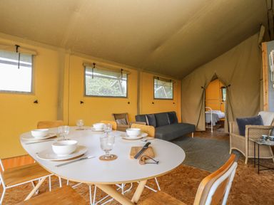 A dining and living area with a white dining table set with bowls glasses and cutlery wooden chairs a gray sofa a wicker chair with cushions and an adjacent bedroom with a white bed frame at Honey @ Stars & Embers Glamping in Grampound