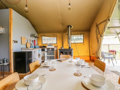 A dining table set with bowls glasses and chairs inside a glamping tent with a stove kitchen area and outdoor deck at Honey @ Stars & Embers Glamping in Grampound