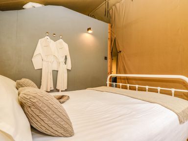 A bedroom with a white bed frame beige pillows white robes hanging on the wall and beige curtains at Honey @ Stars & Embers Glamping in Grampound