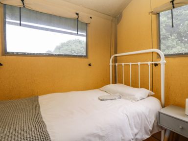 A bed with white metal frame next to a nightstand in a room with yellow fabric walls and two windows at Honey @ Stars & Embers Glamping in Grampound
