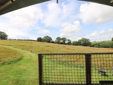 A grassy field with a walking path and trees in the distance seen from behind a netted fence with a fire pit on the right at Honey @ Stars & Embers Glamping in Grampound