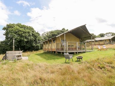 A raised tent structure with a covered porch and outdoor furniture on a grassy area near wooden hot tub and fire pit at Bumble @ Stars & Embers Glamping in Grampound