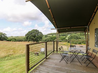 A wooden deck with a table and chairs overlooking a grassy field and trees at Bumble @ Stars & Embers Glamping in Grampound