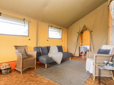 An interior of a tent with a sofa, two wicker chairs, a small table with cups and magazines, and a view of a bedroom with a bed at Bumble @ Stars & Embers Glamping in Grampound