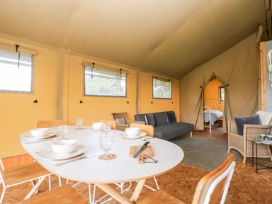 A dining table set with bowls and glasses inside a tent with chairs, sofa, and a bed visible through an open doorway at Bumble @ Stars & Embers Glamping in Grampound