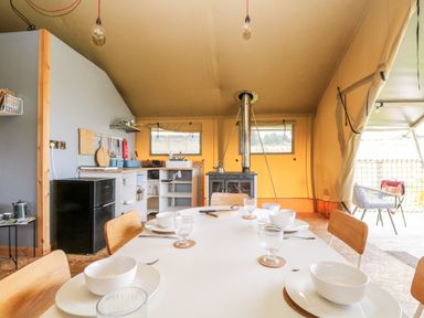 A dining area with a white table set for four with bowls and glasses next to a kitchen with a black refrigerator and wood stove at Bumble @ Stars & Embers Glamping in Grampound