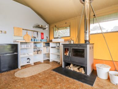 A kitchen area with a black stove and wood storage next to open shelving with kitchenware and a small fridge at Bumble @ Stars & Embers Glamping Grampound
