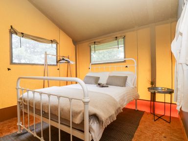 A bedroom with a white metal bed frame a beige blanket two gray pillows a wooden clothing rack with hangers and a small round side table at Bumble @ Stars & Embers Glamping in Grampound