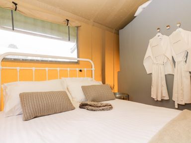 A bedroom with a white metal bed frame two pillows beige cushions two white robes hanging on hooks and a small table at Bumble @ Stars & Embers Glamping in Grampound
