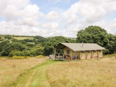 A tent structure with a covered porch and chairs in a grassy field with trees and hills in the background at Bumble @ Stars & Embers Glamping in Grampound