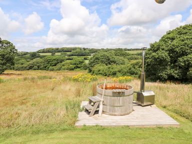 A wooden hot tub on a deck in a grassy field with trees and hills in the background at Bumble @ Stars & Embers Glamping in Grampound