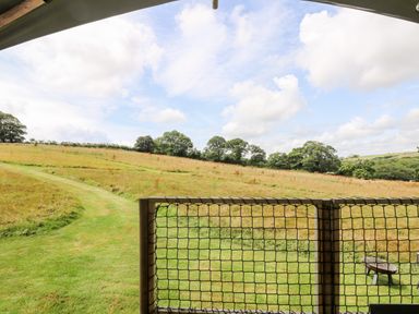 A view of a grassy field with trees and a curved path from behind a netted wooden railing at Bumble @ Stars & Embers Glamping in Grampound