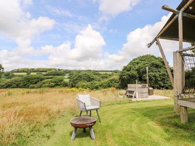 An outdoor area with a chair, a fire pit, a wooden hot tub, and a grassy field with trees in the background at Bumble @ Stars & Embers Glamping in Grampound
