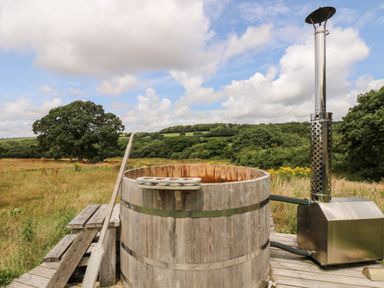 An outdoor wooden hot tub with a metal stove and chimney in a grassy field with trees and hills at Bumble @ Stars & Embers Glamping in Grampound