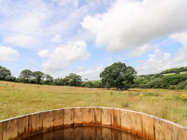 A wooden hot tub in a field with trees and hills in the background at Bumble @ Stars & Embers Glamping Grampound
