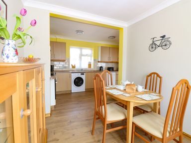 A dining area with four wooden chairs and a table set with teapot and bread next to a kitchen with wooden cabinets and washing machine at Lillies Pad in Methwold near Feltwell