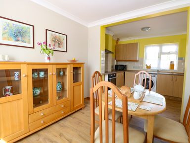 A dining area with a wooden table and chairs adjacent to a kitchen with wooden cabinets and a glass-fronted cabinet with decorative items at Lillies Pad Methwold near Feltwell