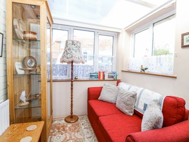 A conservatory with a red sofa and gray cushions a wooden display cabinet with decorative items and a floral floor lamp at Lillies Pad Methwold near Feltwell