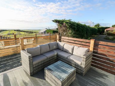 An outdoor seating area with a gray cushioned sectional sofa and a matching glass-top coffee table on a wooden deck at Caban Tywod in Abersoch