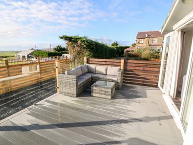 An outdoor patio with a gray wicker corner sofa and glass top table at Caban Tywod in Abersoch