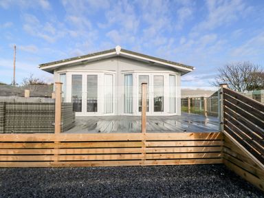 A raised wooden deck with glass railings and a white building with double doors at Caban Tywod in Abersoch