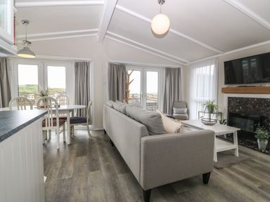 An open living room and dining area with a gray sofa a white dining table with chairs a fireplace and a wall-mounted television at Caban Tywod in Abersoch