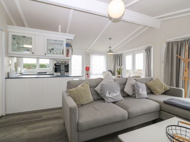 A living room with a grey sofa and cushions facing an open kitchen with glass cabinets and a dining table near windows at Caban Tywod in Abersoch