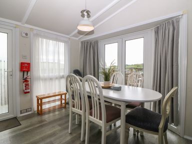 A dining room with white oval table six chairs a plant on the table a window and glass doors with curtains at Caban Tywod in Abersoch