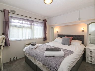 A bedroom with a double bed, pillows, folded towels, built-in cabinets, bedside tables, a window with curtains and a ceiling light at Caban Tywod in Abersoch