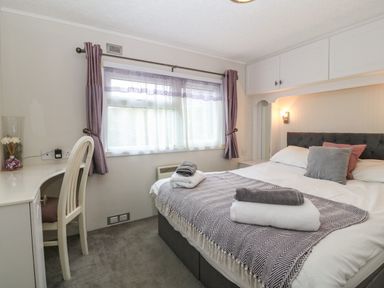 A bedroom with a bed towels on it a desk chair window with curtains and overhead cupboards at Caban Tywod in Abersoch