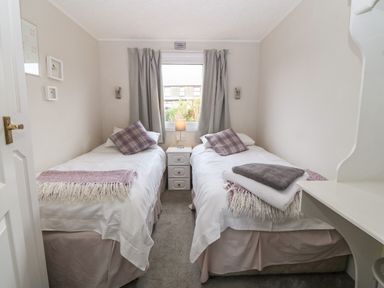 A bedroom with two single beds with pillows and throws a nightstand with a lamp between the beds and a window with curtains behind the beds at Caban Tywod in Abersoch