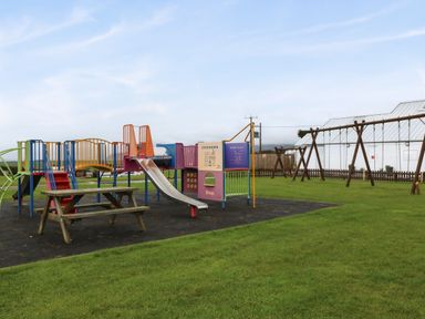An outdoor playground with a slide climbing structures and swings on grass at Caban Tywod in Abersoch