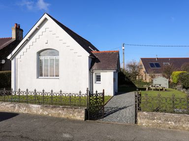 A white building with a black roof and metal fence with a garden bench and shed behind it at Capel Hermon in Llanynghenedl near Bodedern