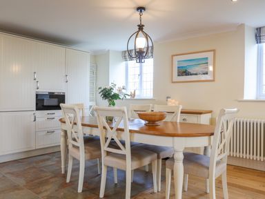 A dining area with a wooden table and six white chairs next to white cabinets and two windows at Capel Hermon in Llanynghenedl near Bodedern