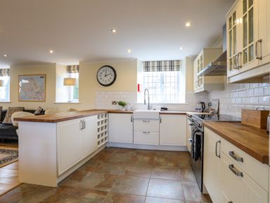 A kitchen with white cabinets wooden countertops a large clock above the sink and windows with blinds at Capel Hermon in Llanynghenedl near Bodedern