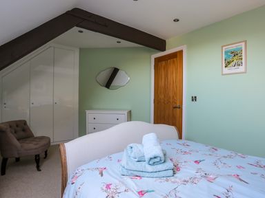 A bedroom with a bed covered in floral bedding and folded towels on top a brown armchair white dresser white built-in closet wooden door and green walls at Capel Hermon in Llanynghenedl near Bodedern