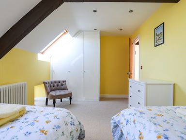 A bedroom with two single beds with floral bedding a tufted chair built in white cupboards a white chest of drawers and yellow walls at Capel Hermon in Llanynghenedl near Bodedern