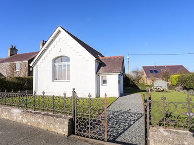A white house with a pitched roof and a gravel driveway with a metal gate in front at Capel Hermon in Llanynghenedl near Bodedern