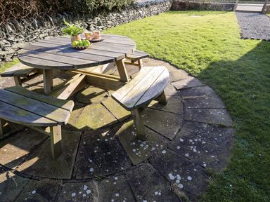 A wooden picnic table with benches on circular stone paving with grass and a stone wall at Capel Hermon in Llanynghenedl near Bodedern