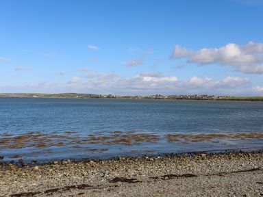 A rocky shoreline with water and a distant view of houses and fields at Capel Hermon in Llanynghenedl near Bodedern