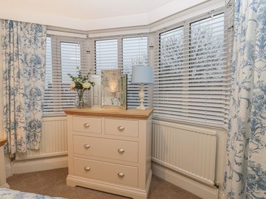 A corner bedroom with a dresser holding a vase of flowers a table lamp and a mirror with white blinds on three windows and blue and white patterned curtains at Hillcrest in Scarborough