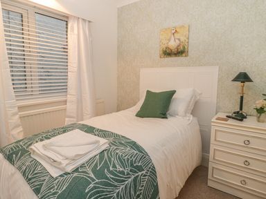 A bedroom with a single bed a green pillow and a folded towel on the bed a window with blinds and curtains a bedside table with a lamp and a vase with flowers and a painting of a duck on the wall at Hillcrest in Scarborough