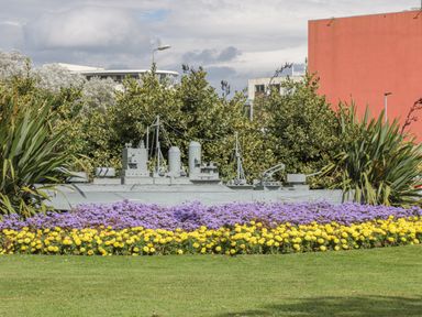 A garden with yellow and purple flowers and a gray model ship surrounded by bushes and buildings at Hillcrest in Scarborough