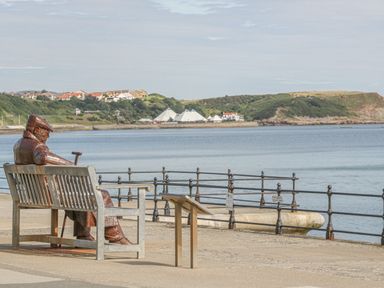 A metal statue of a seated man with a cane on a bench overlooking water with a railing and green hills in the background at Hillcrest in Scarborough